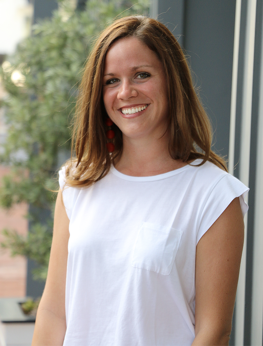 A woman with long light brown hair wearing a white sleeveless top and red earrings, smiling in an outdoor setting with greenery in the background.