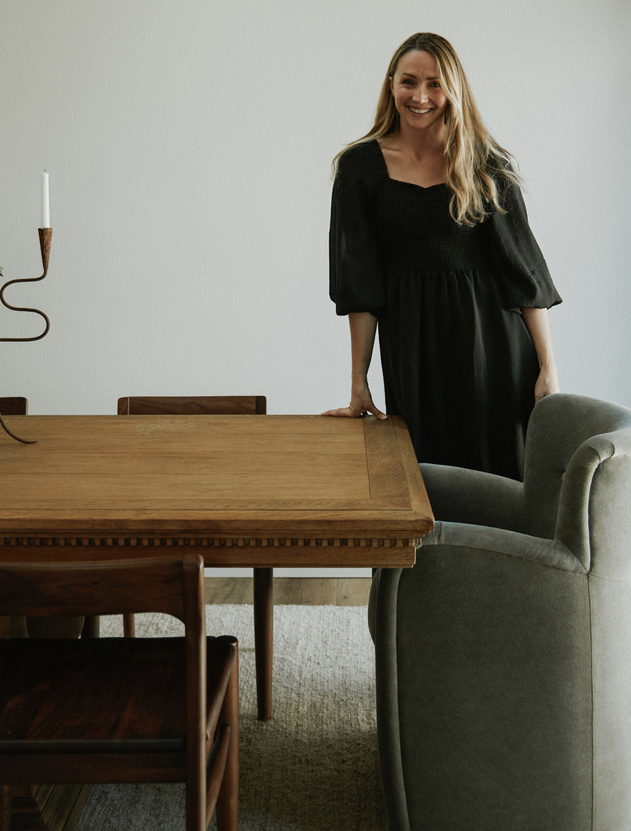 A woman with long blonde hair, wearing a black dress, standing and smiling by a wooden dining table with a candle holder, next to a green armchair.