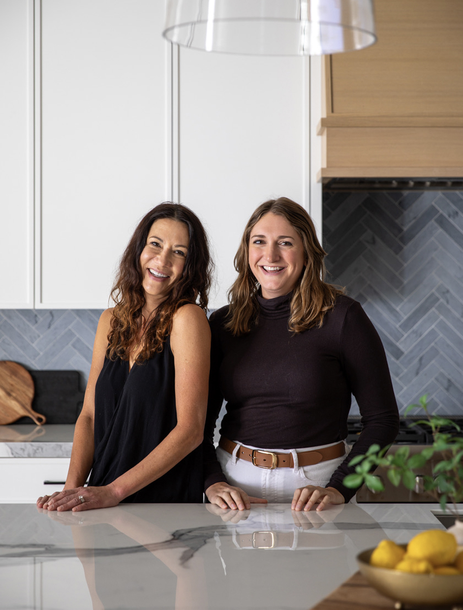 Two women smiling, one with long dark hair in a black sleeveless top, the other with light brown hair in a black long-sleeve top, standing in a modern kitchen.