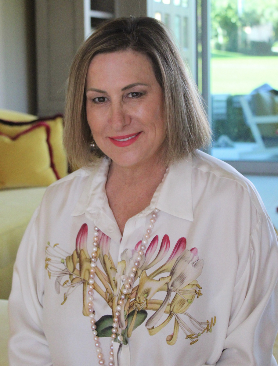 A woman with short blonde hair, wearing a floral print blouse and a pearl necklace, smiles warmly in a cozy, well-lit living room.