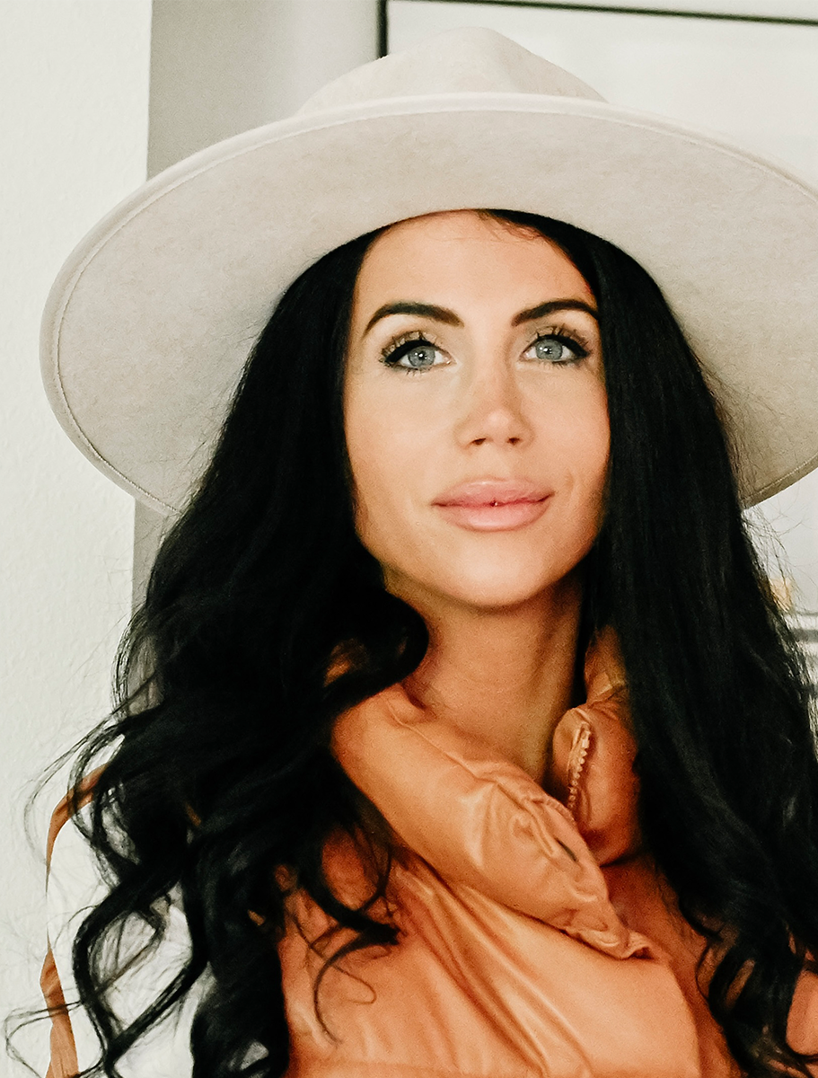 A portrait of a woman with long dark hair, wearing a beige hat and an orange puffy vest, looking directly at the camera with a neutral background.