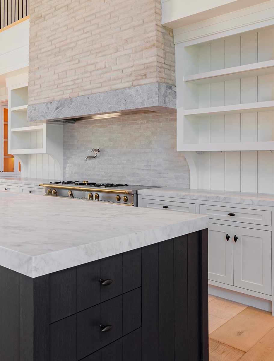 Modern kitchen with a black island and white marble countertop, complemented by light gray cabinets and a brick range hood, offering a blend of classic and contemporary design elements.