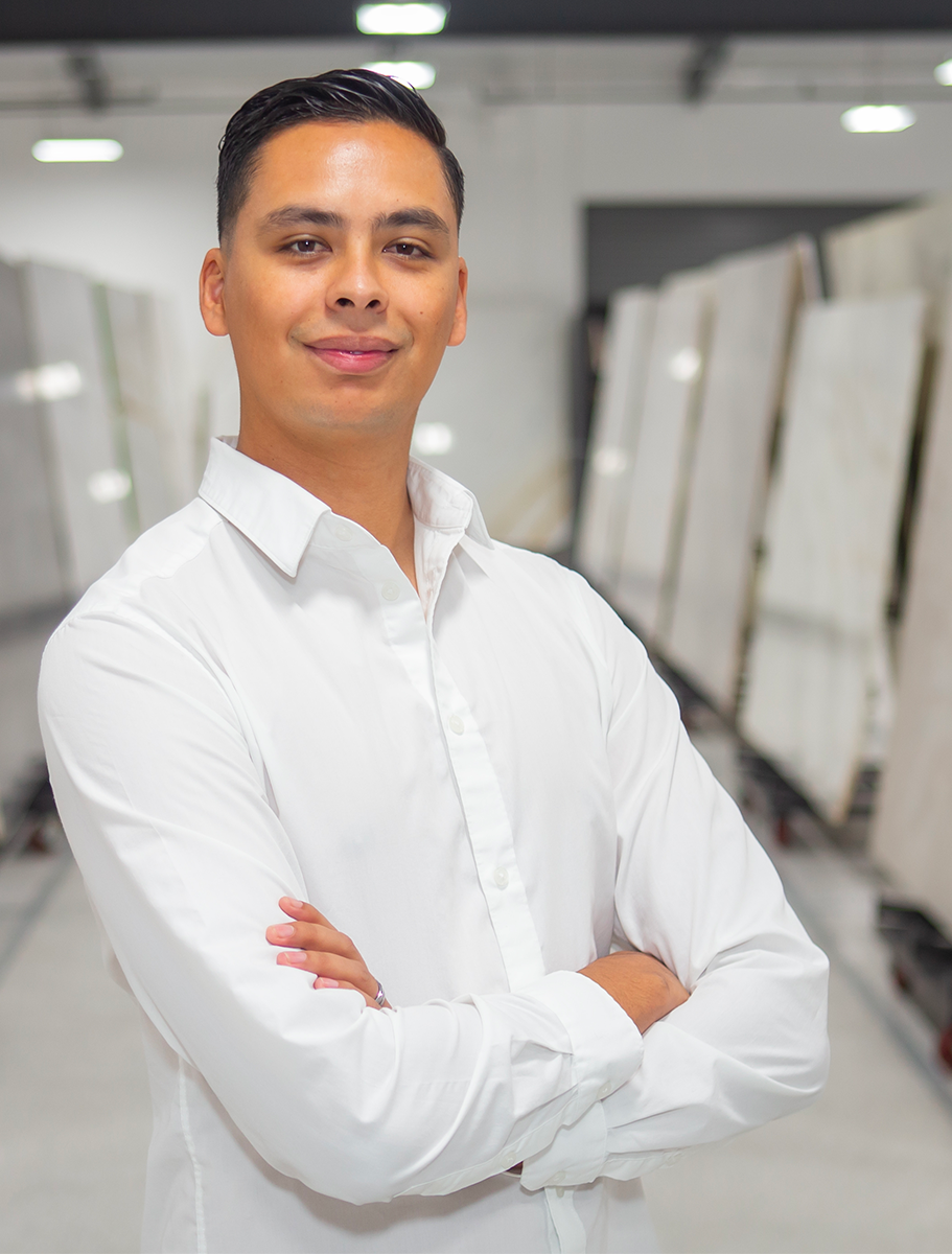 A confident man in a white shirt standing with folded arms in a bright, modern setting, surrounded by rows of marble slabs in the background.