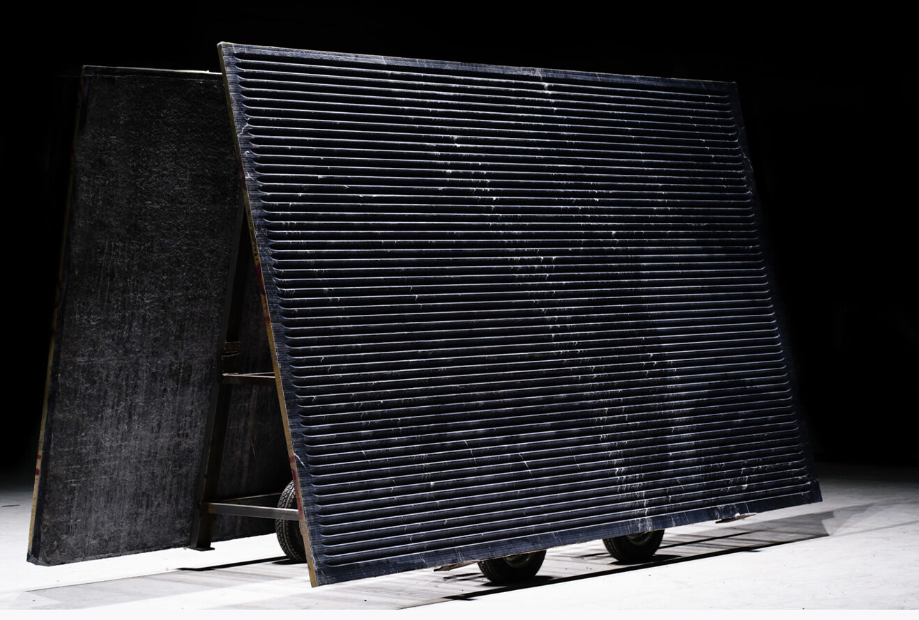 The image depicts a large, tilted black panel with horizontal grooves, mounted on a wheeled frame, set against a dark background in a studio.