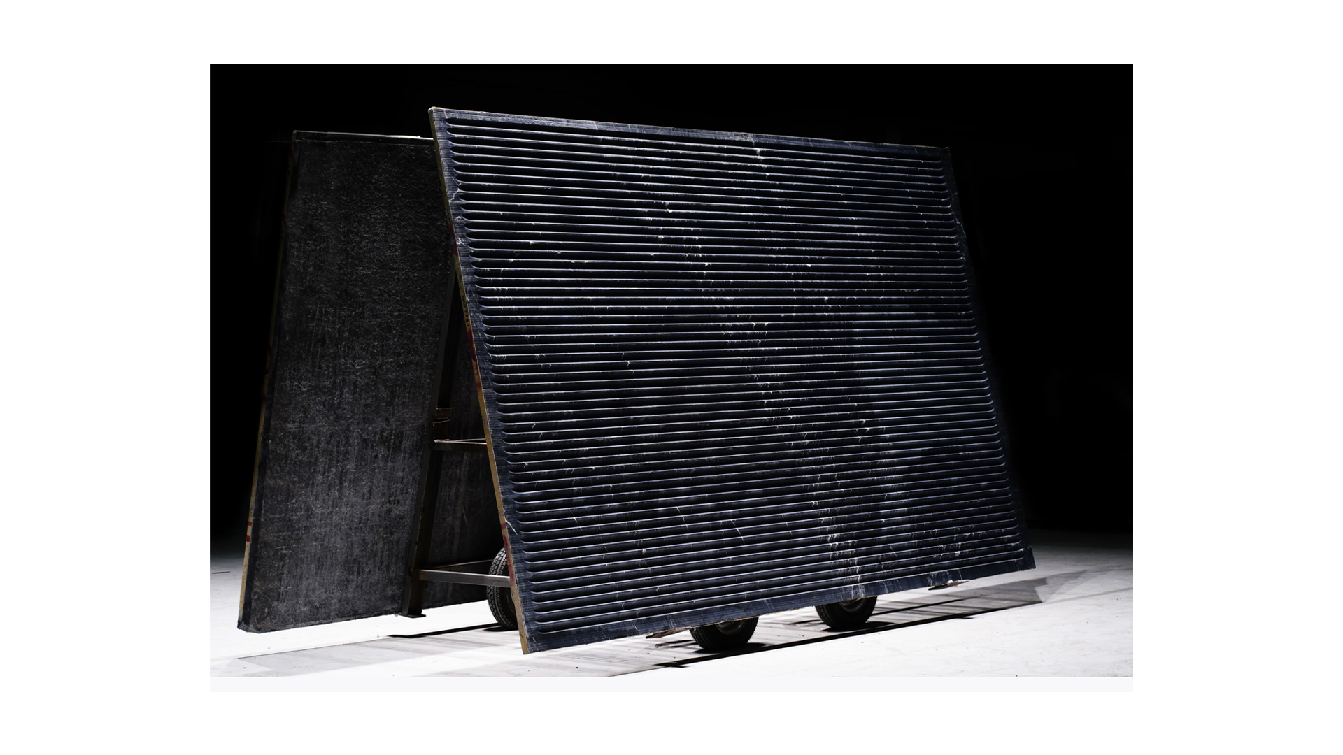 The image depicts a large, tilted black panel with horizontal grooves, mounted on a wheeled frame, set against a dark background in a studio.