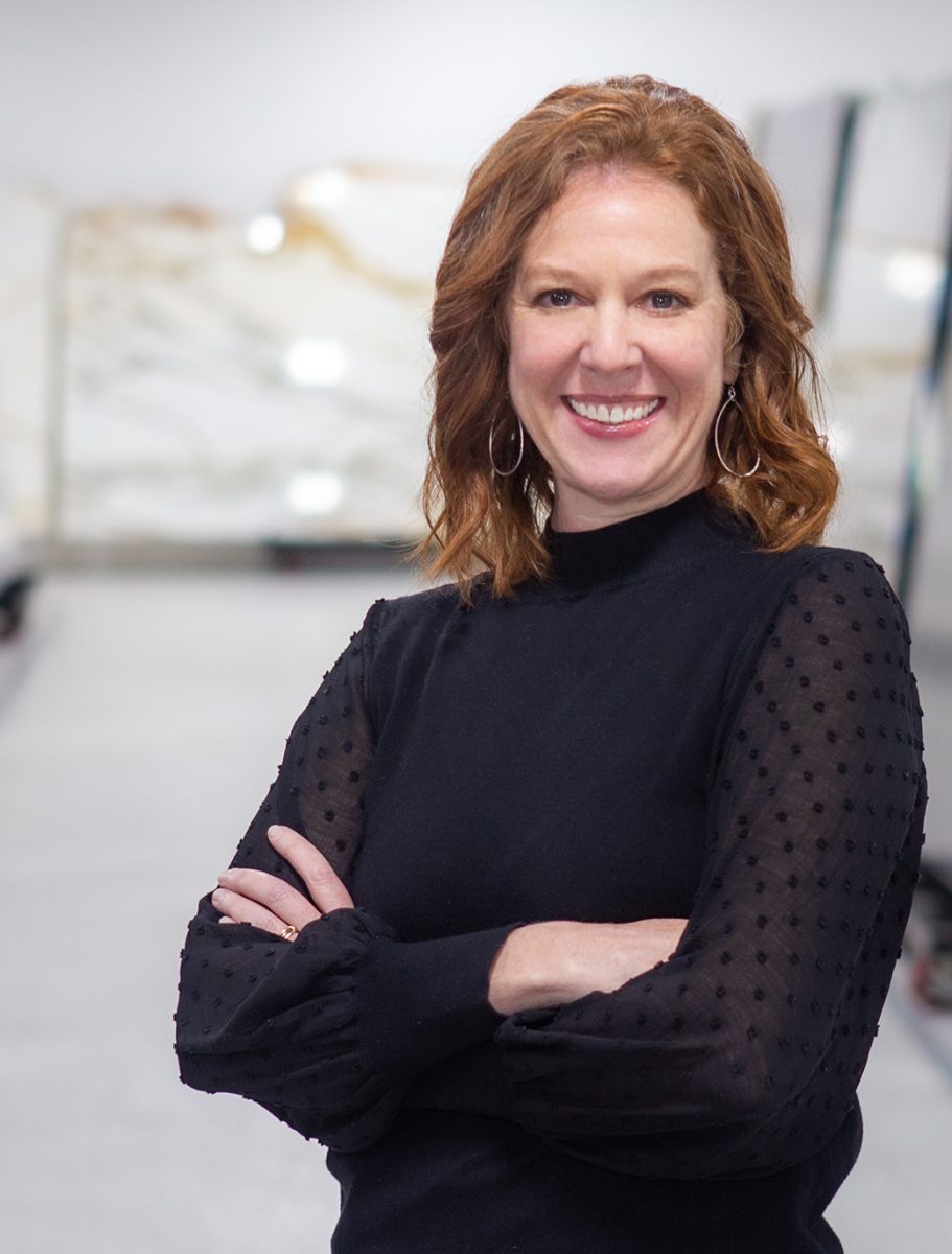 A woman with wavy red hair, wearing a black top with sheer, dotted sleeves, stands confidently with arms crossed in front of large stone slabs in a bright indoor space.